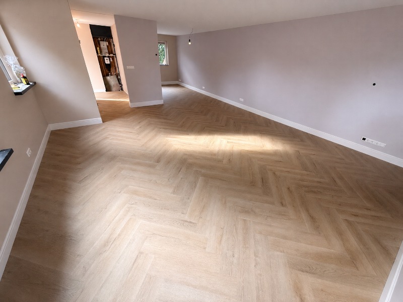 Woonkamer met een lichte, houtlook PVC-plakvloer in visgraatpatroon en grijsgeschilderde wanden.