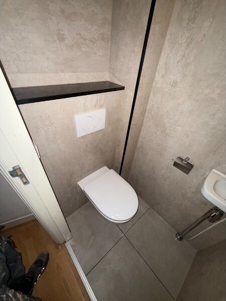 Afgewerkt hangend toilet met betonlook pvc-wandpanelen, een zwarte legplank en een fonteintje.