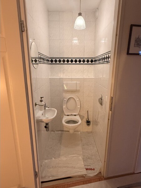 Verouderd toilet op de begane grond met witte tegels en een zwart-witte diamantrand.