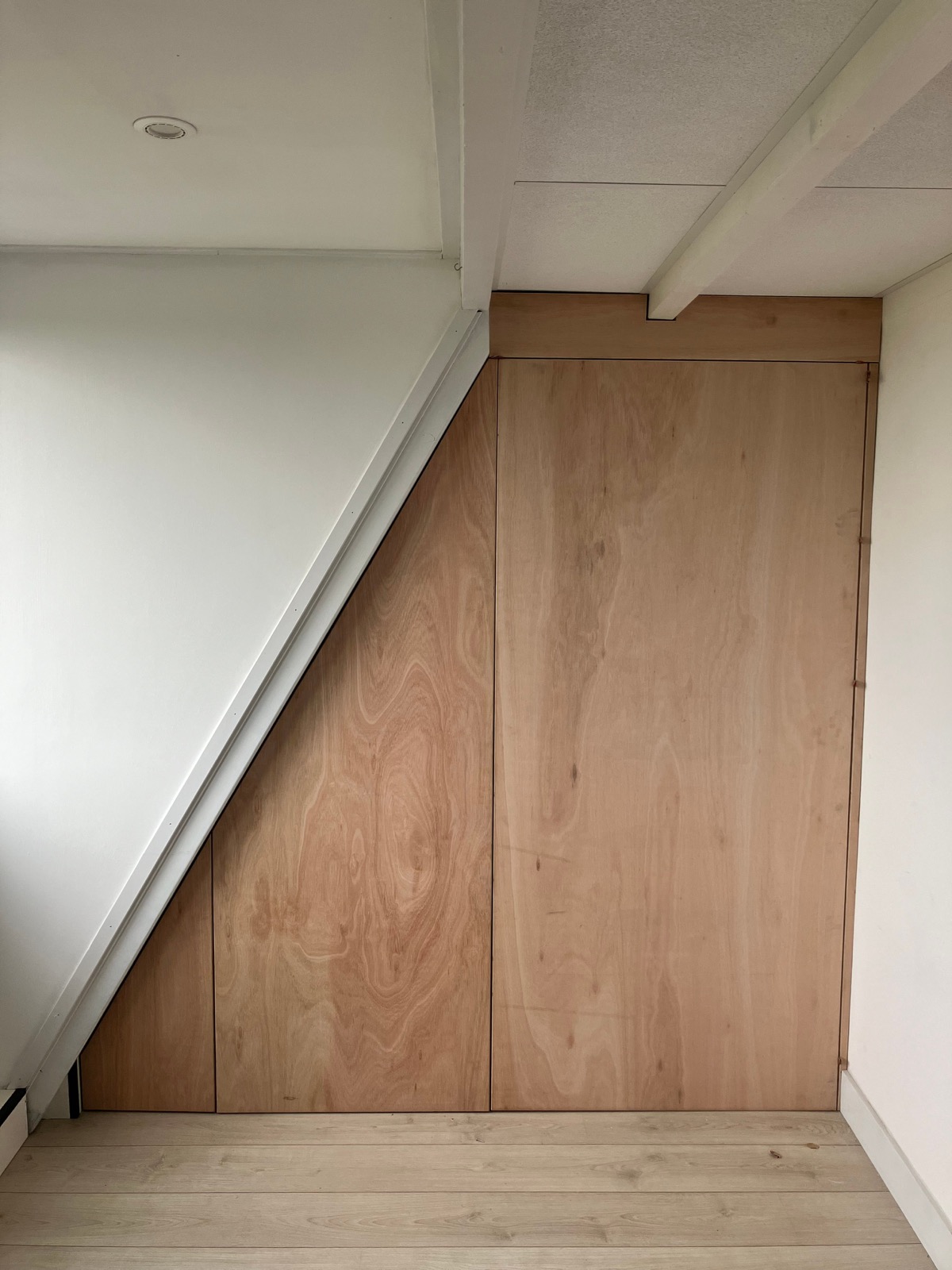 Gesloten kastenwand in licht multiplex tegen een schuine wand, met deuren.
