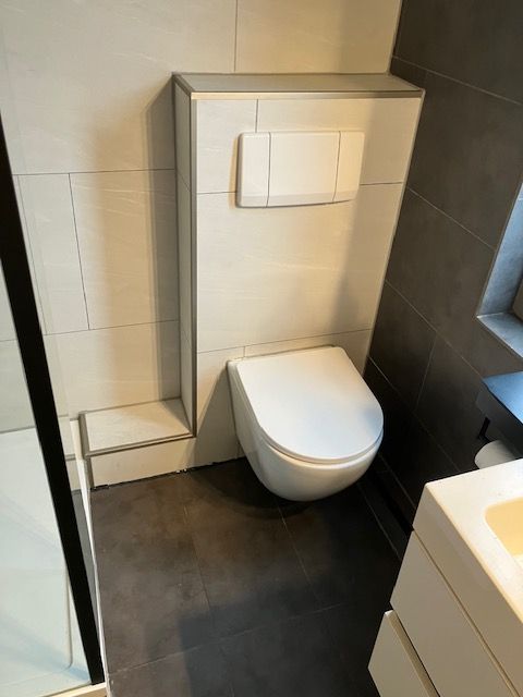 Toiletombouw afgewerkt met crèmekleurige tegels, spoelplaat en donkere vloertegels.