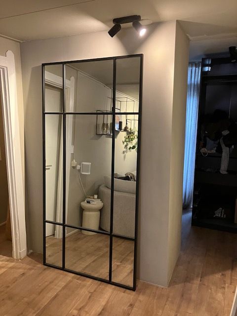 Grijs geschilderde wand met een grote spiegel in een stalen frame en een spot erboven.