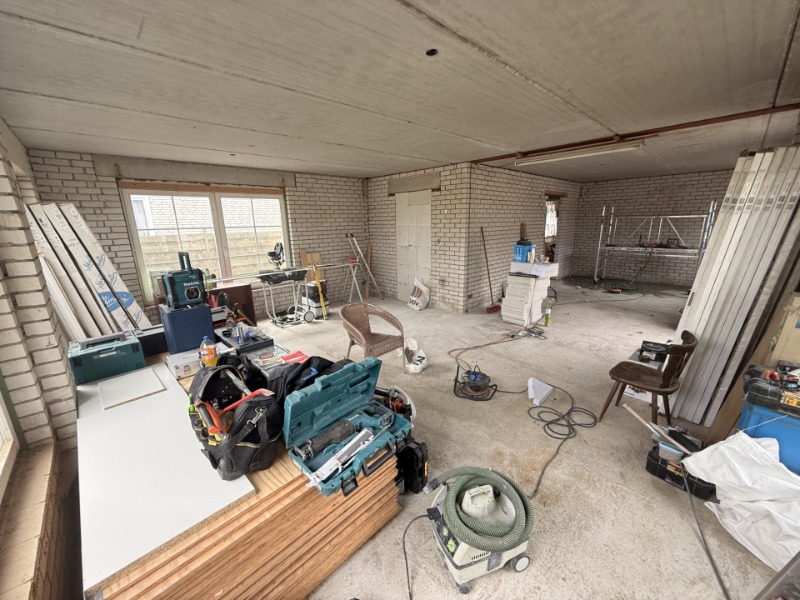 Kamer in aanbouw met witgeschilderde bakstenen wanden, stapels gereedschapskisten en bouwmateriaal.